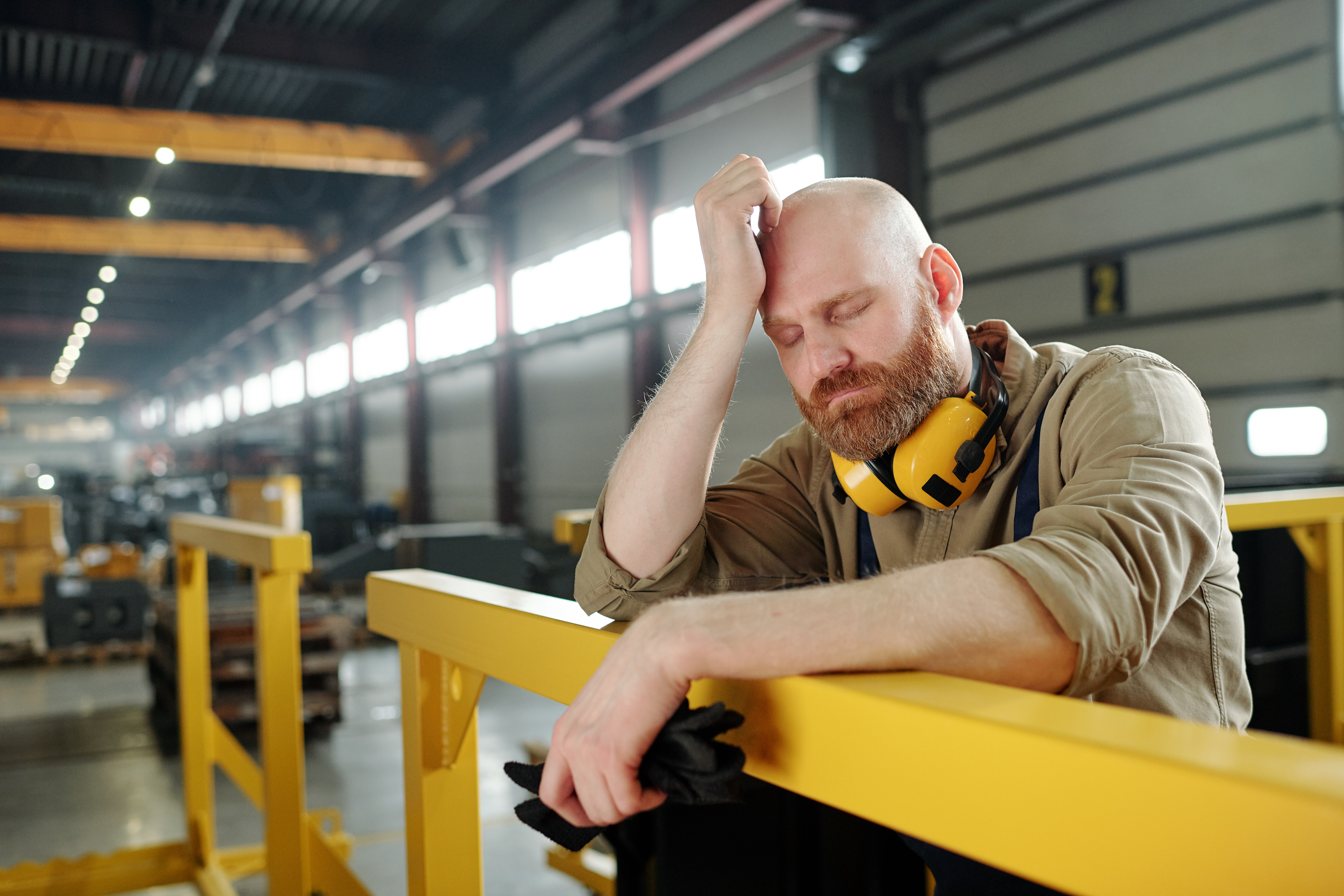 Tired or sick bald engineer touching his head while leaning by bar at break in the middle of working day in workshop
